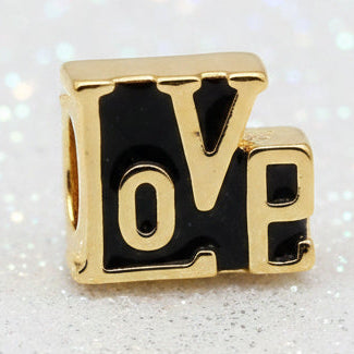 A square charm featuring the word 'LOVE' in raised gold letters with a glossy black enamel background, shown on a dark reflective surface.