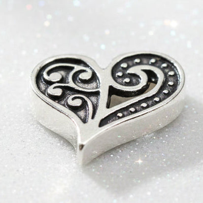 A silver-toned heart-shaped charm with intricate filigree cutouts and small dot accents, shown on a dark reflective background.