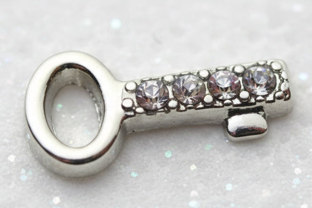 A silver-toned key-shaped charm or pendant featuring a row of clear rhinestones on the handle, set against a light gray textured background.