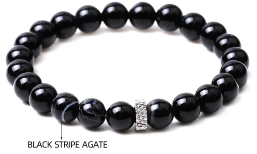 A black beaded bracelet with a small silver rhinestone accent, resting on a white stone with a beach and turquoise ocean in the background.