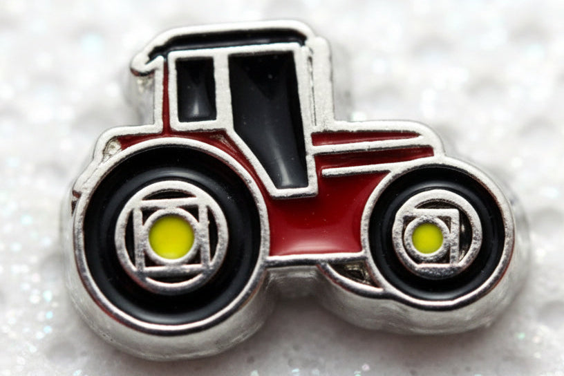 A small tractor-shaped charm or bead with a silver-toned outline, black wheels and cabin, a red body, and bright yellow-green center details, shown on a white textured background.