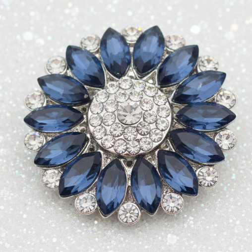 A round floral snap button featuring a silver-tone setting with a central circle of clear crystals and outer petals of faceted dark blue marquise-shaped crystals.