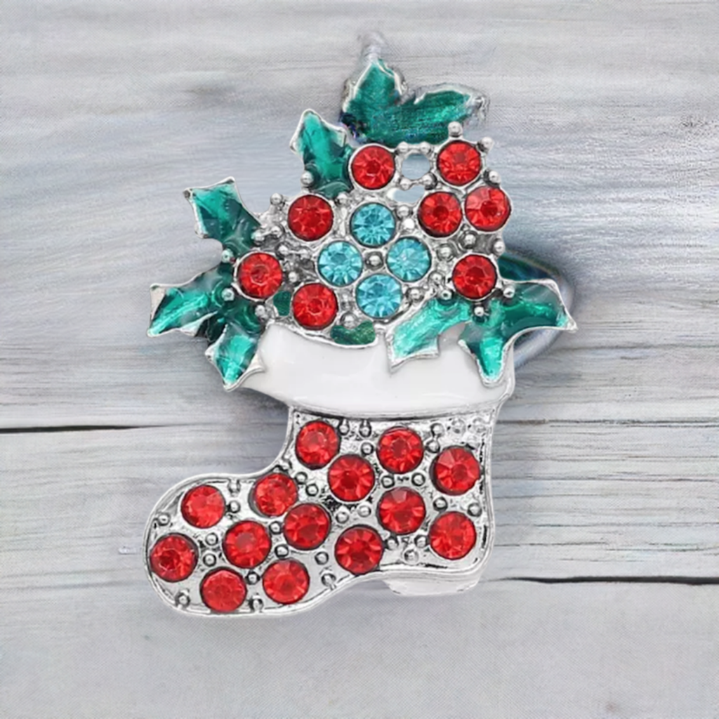 A Christmas stocking snap button with a silver-tone outline, decorated with red and light blue rhinestones and green holly leaves, shown on a light gray wood background.
