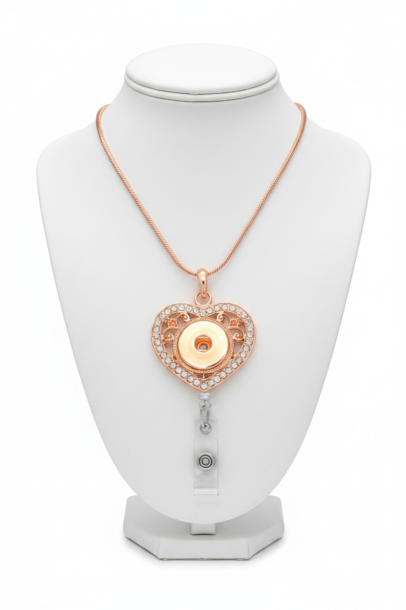 A rose gold heart-shaped pendant necklace with a clear rhinestone border and a central gold-tone snap button, attached to a silver retractable badge reel on a white background.