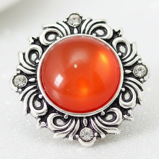 An ornate silver snap button with a large round orange-red cabochon gemstone and small clear stones, displayed on a white background.