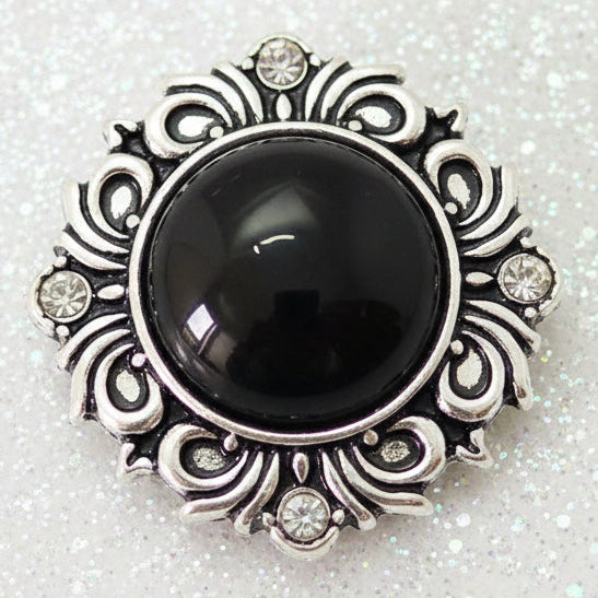 A vintage-style silver-tone snap button featuring a large, smooth black oval cabochon stone in the center, surrounded by an ornate filigree border with small clear rhinestones.