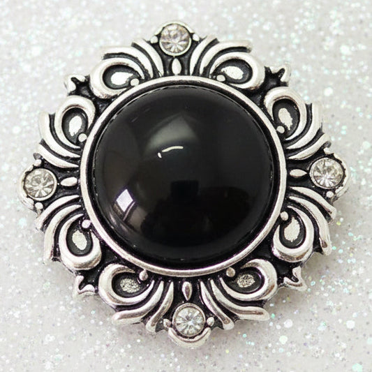 A vintage-style silver-tone snap button featuring a large, smooth black oval cabochon stone in the center, surrounded by an ornate filigree border with small clear rhinestones.