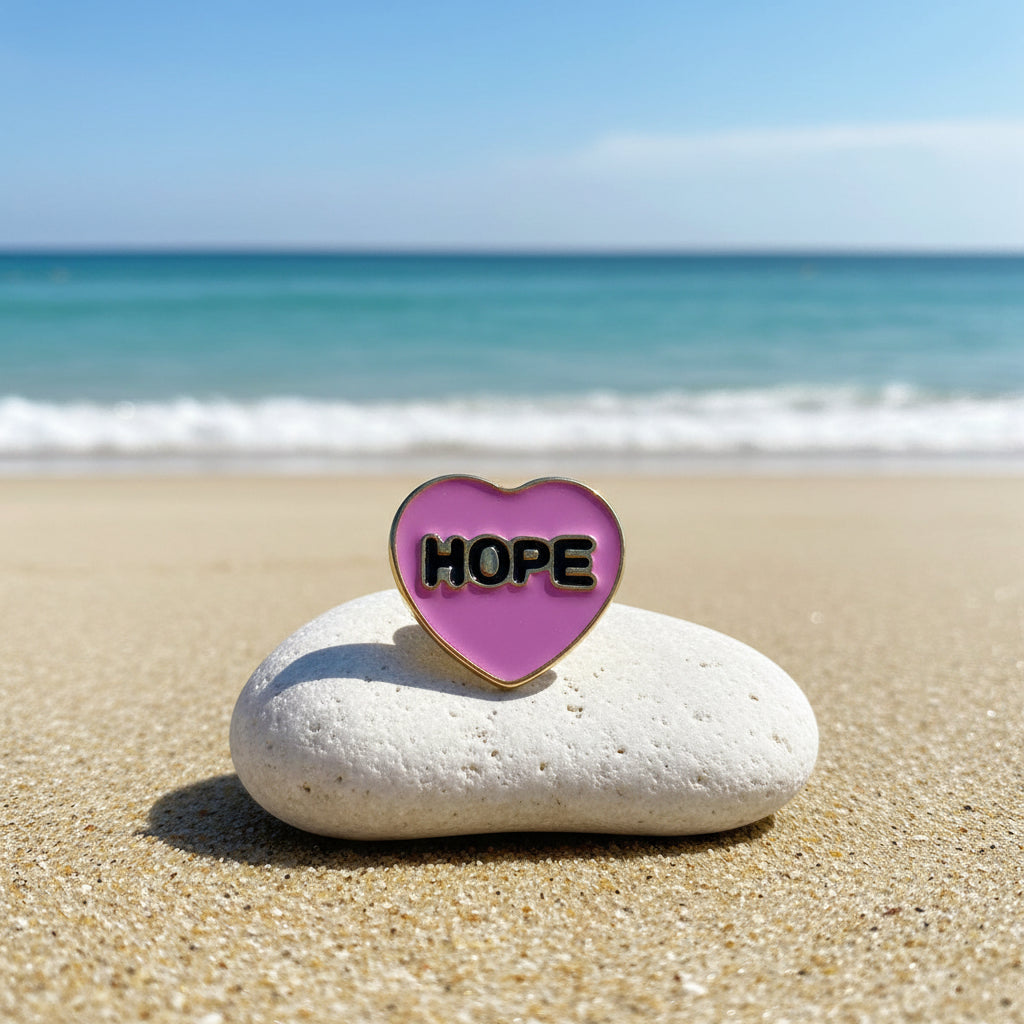 A heart-shaped enamel snap button with a pink center, gold border, and the word 'HOPE' in black letters with gold outline.