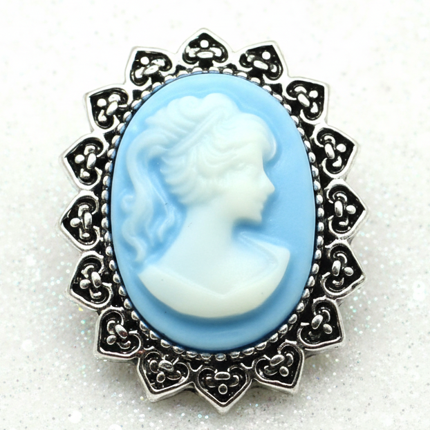 A vintage-style cameo snap button with a light blue and white profile design in a silver-toned filigree frame, displayed on a white backdrop