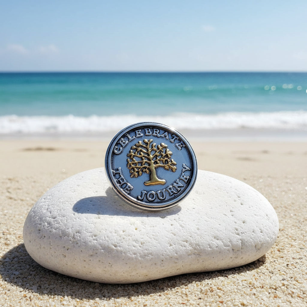 A round silver-toned snap button with a gold-toned tree design and the words 'CELEBRATE BRAVE THE JOURNEY' around the edge, resting on a sparkly white surface.