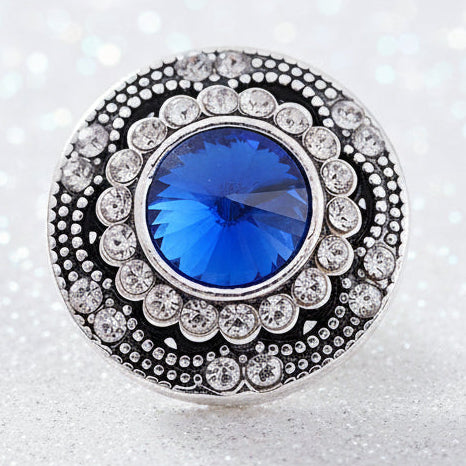 A silver-toned snap button with a large round blue center stone and surrounding clear stones, displayed on a smooth white rock on a sparkly white backdrop