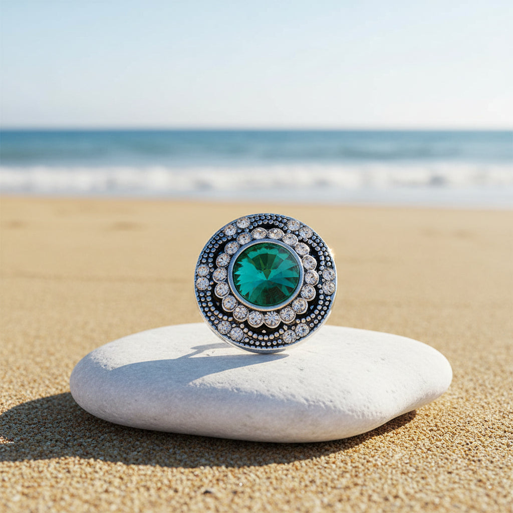 A round silver-tone snap button featuring a large, vibrant green faceted crystal in the center, surrounded by a double ring of small clear rhinestones on a sunny beach