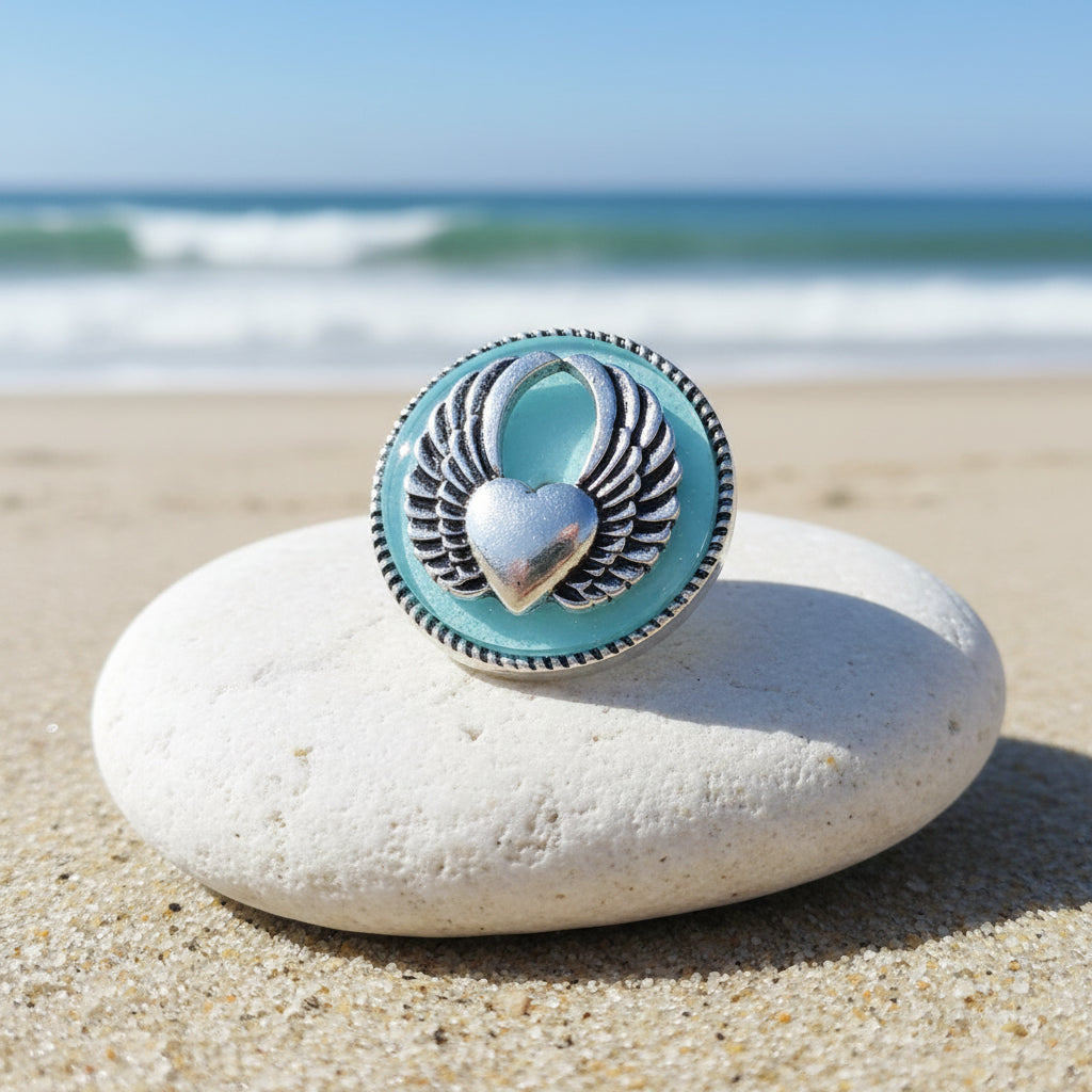 A round light blue snap button featuring a silver-toned metal angel wing design with a central heart charm, finished with a textured beaded border.