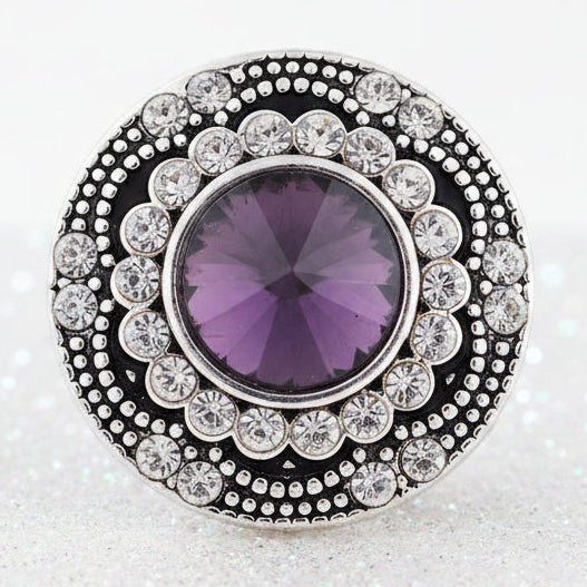 A round silver snap button with a large purple faceted center stone and a halo of small clear stones, displayed on a smooth white stone on a sandy beach with the ocean in the background.