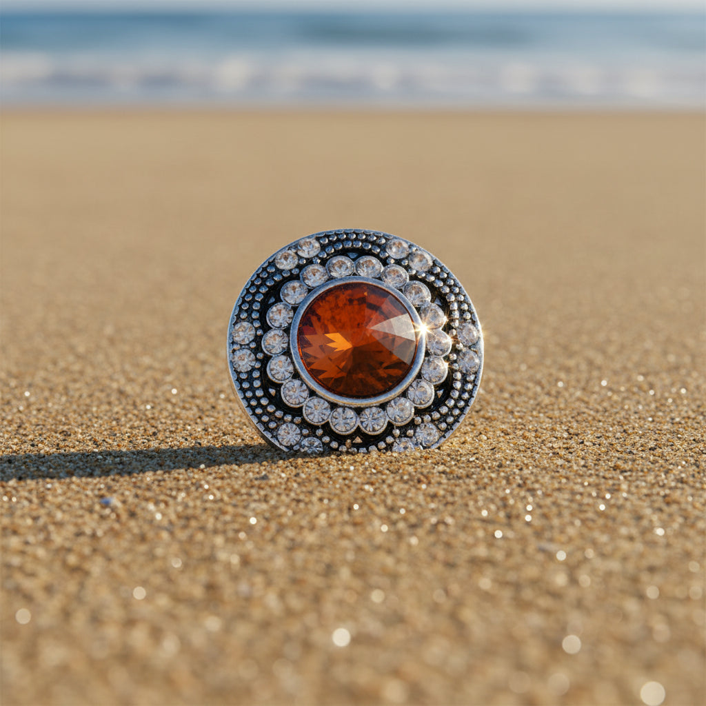 A round silver-toned snap button embellishment featuring a large faceted orange center stone surrounded by a halo of small clear rhinestones on a beach