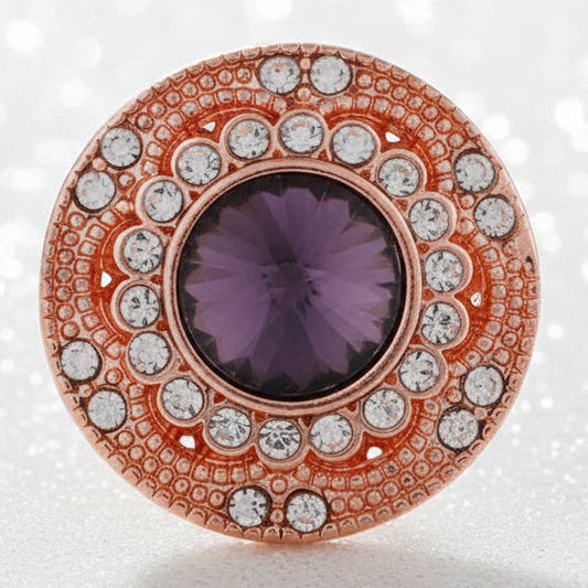 A rose gold snap button with a purple amethyst center and clear accent stones, resting on a smooth white stone on a sandy beach with the ocean in the background.