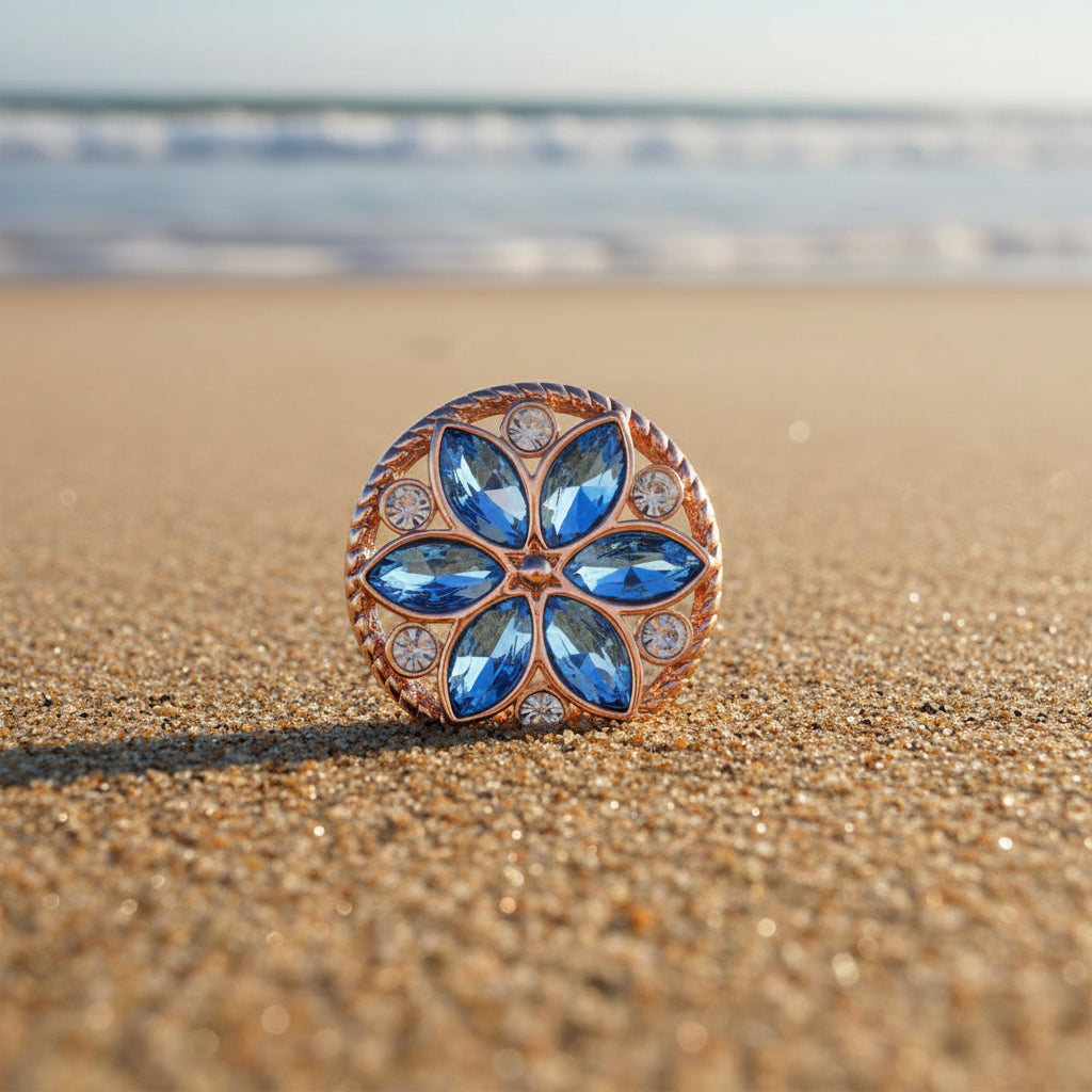 A round rose gold snap button featuring a floral design with blue marquise-cut stones and small clear accent stones, set against a sparkly white background.