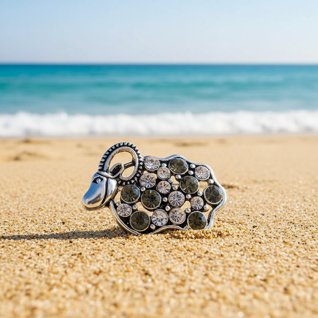 A silver-toned sheep-shaped snap button embellished with round dark gray and clear rhinestones, shown on a beach background.