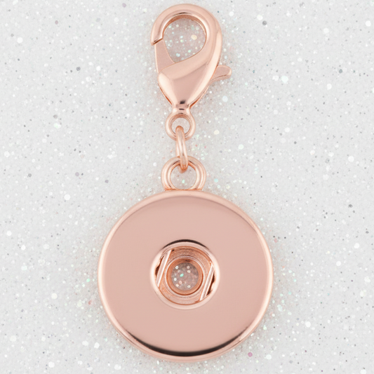A rose gold snap button badge with a lobster clasp, shown on a light gray glittery background.