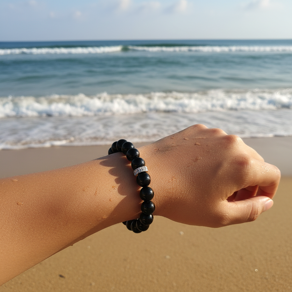 A black beaded bracelet with a silver-toned textured accent bead, worn by a model on a sandy beach with the ocean waves blurred in the background.