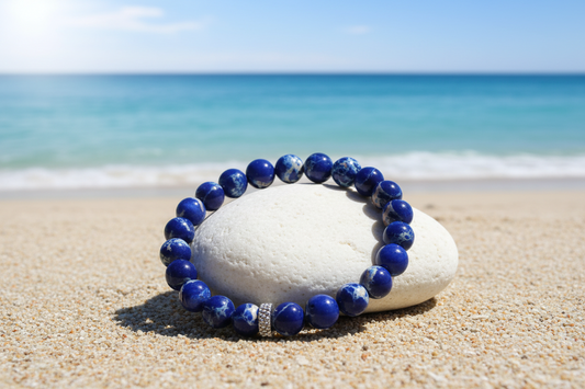 A beaded stretch bracelet made of deep blue lapis lazuli stones with a small silver spacer, resting on a smooth white stone in sandy beach with the ocean in the background.