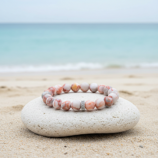 A beaded stretch bracelet with round pink, white, and gray marbled beads and a small silver rhinestone accent, displayed on a smooth white stone on a sandy beach with the ocean in the background.