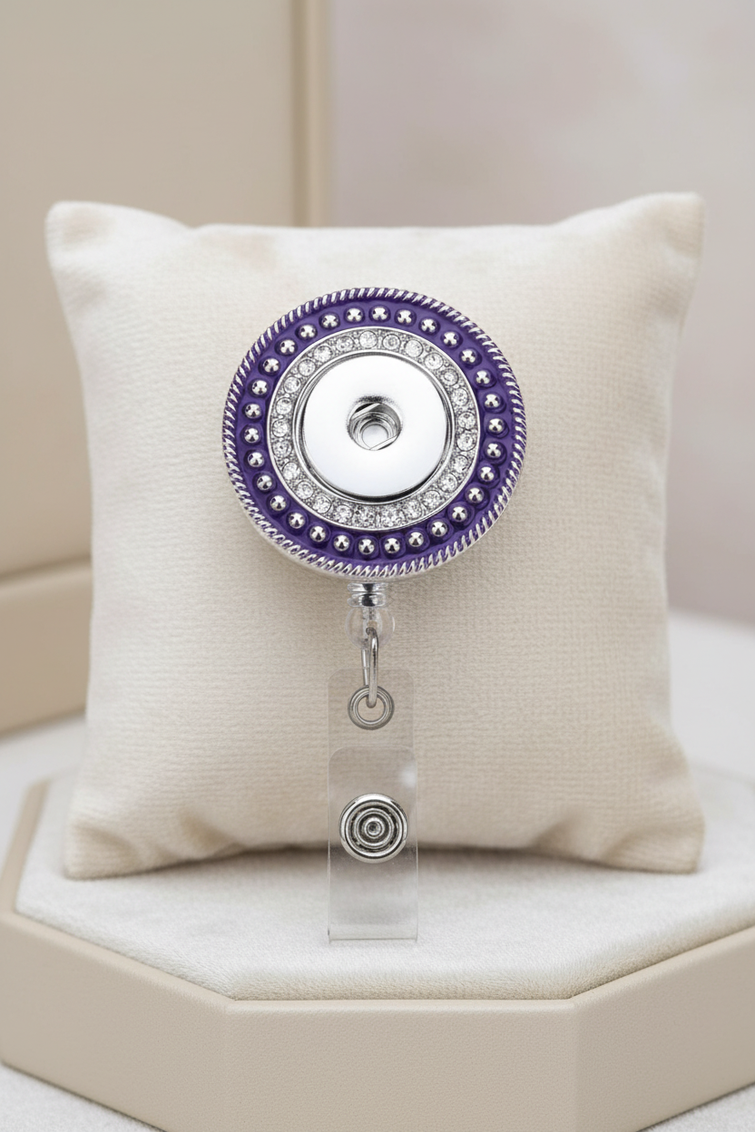 A circular retractable badge reel with a purple and silver design, featuring a clear plastic clip at the bottom.