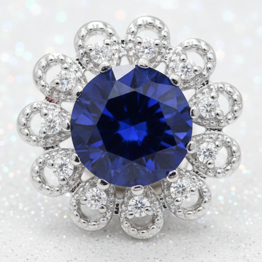 A floral cluster snap button featuring a large round brilliant-cut deep blue sapphire center stone surrounded by a halo of small clear stones in a silver-toned setting, isolated on a white background.