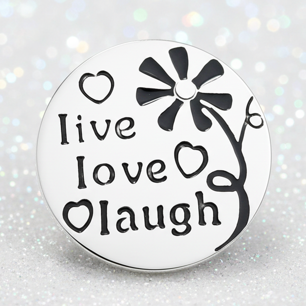 A snap button with black text reading 'live love laugh' and a flower design, resting on a smooth white stone on a sandy beach with the ocean in the background.