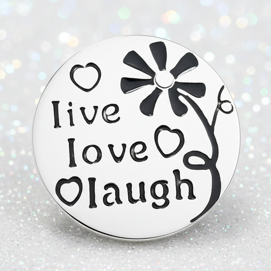A snap button with black text reading 'live love laugh' and a flower design, resting on a smooth white stone on a sandy beach with the ocean in the background.