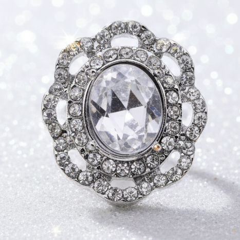 A silver-toned snap button with a large oval clear gemstone and surrounding clear stones, displayed on a smooth white stone at the beach with the ocean and sky in the background.