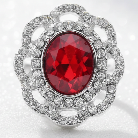 A silver-toned snap button with a large oval red gemstone and surrounding clear stones, displayed on a smooth white stone in a sandy beach setting with turquoise water in the background.
