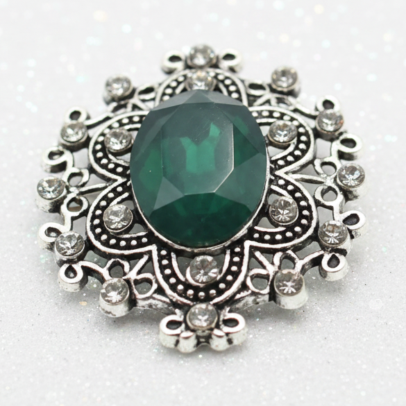 An ornate silver snap button with a large oval green gemstone sits on a smooth white stone placed on sandy beach, with the ocean and sky blurred in the background.