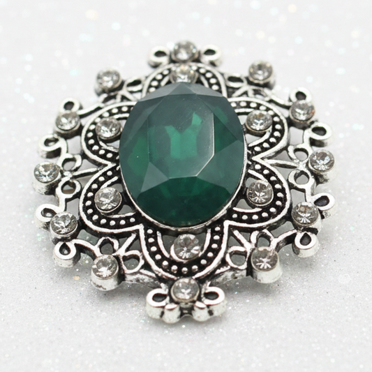 An ornate silver snap button with a large oval green gemstone sits on a smooth white stone placed on sandy beach, with the ocean and sky blurred in the background.