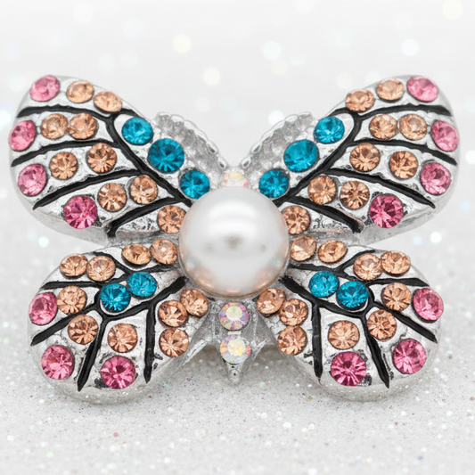 A butterfly-shaped snap button featuring a central white pearl surrounded by pink, peach, and blue rhinestones on a silver-toned setting, photographed on a sparkly white background.