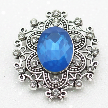 An ornate silver-toned snap button with a large oval blue gemstone and small clear stones, displayed on a smooth white stone at the beach with the ocean and sand blurred in the background.