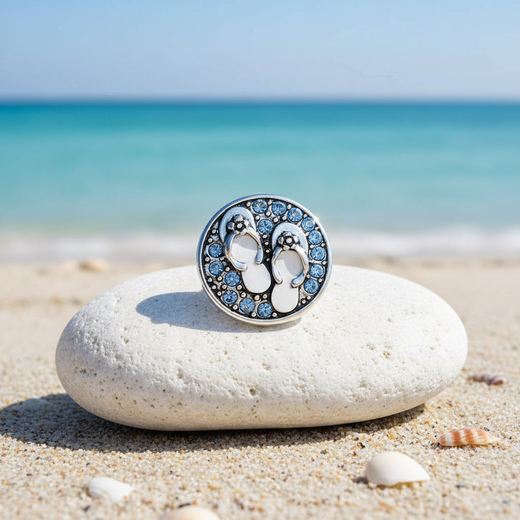 A round silver-toned snap button featuring a pair of flip-flop sandals embellished with light blue rhinestones