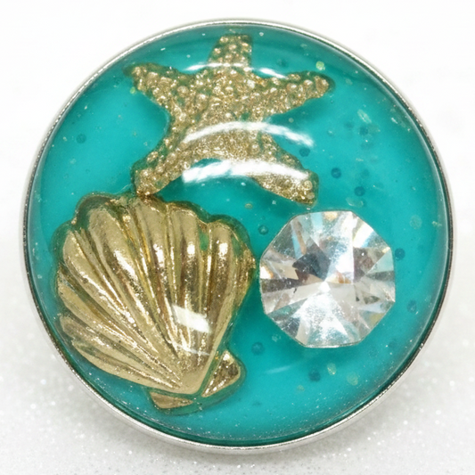 A round turquoise resin snap button  featuring a gold glitter starfish, a gold seashell, and a clear faceted rhinestone, set in a silver-toned metal frame.