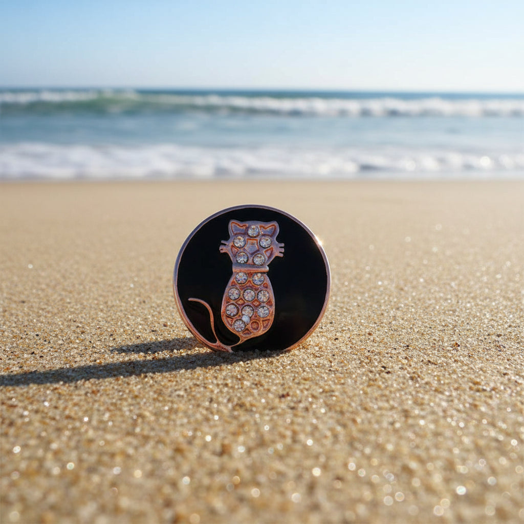A round enamel snap button featuring a rose gold cat silhouette with clear rhinestones on a black background, set against a beach