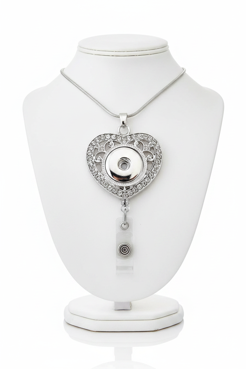 A silver heart-shaped pendant necklace with rhinestone detailing and a clear retractable badge reel attached below, shown on a plain white background.  Pair with statement snaps.