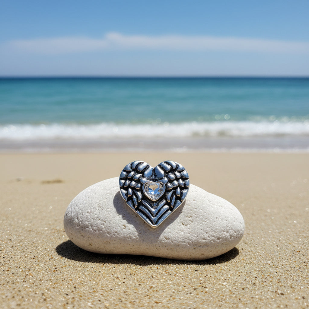 A heart-shaped silver metal snap button with intricate wing-like patterns and a clear rhinestone in the center 