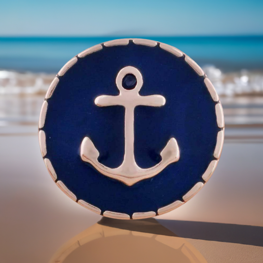 A round navy blue and silver anchor snap button with a scalloped silver border, displayed on a reflective surface with a blurred beach and ocean background.