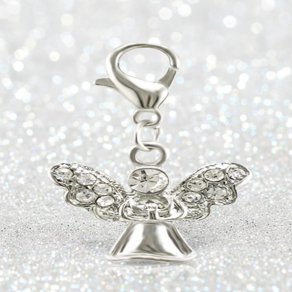 A silver angel charm with clear rhinestone wings and a lobster clasp, displayed on a reflective surface against a soft, blurred background.