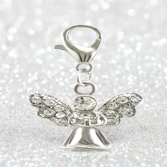 A silver angel charm with clear rhinestone wings and a lobster clasp, displayed on a reflective surface against a soft, blurred background.