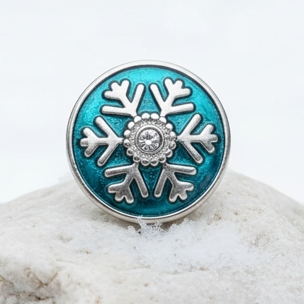 A round silver-toned ring with a turquoise blue enamel snowflake design, resting on a snow-covered rock with a blurred winter forest background.
