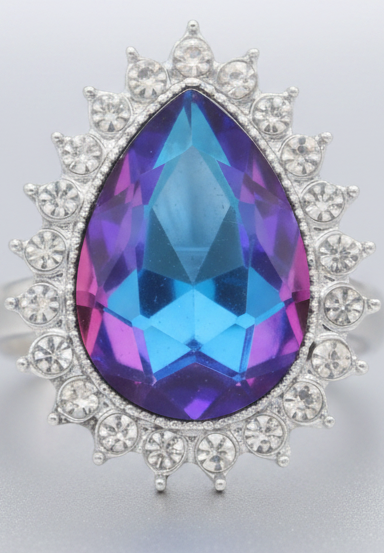 A teardrop-shaped iridescent gemstone snap with a blue, purple, and pink color-shifting center, surrounded by a silver-tone halo of clear cubic zirconia stones, set against a dark green background.