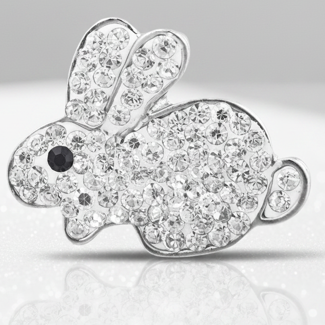 A silver bunny-shaped snap button covered in clear rhinestones, with a small black eye detail, displayed on a white surface with pastel Easter eggs and baskets in the background.