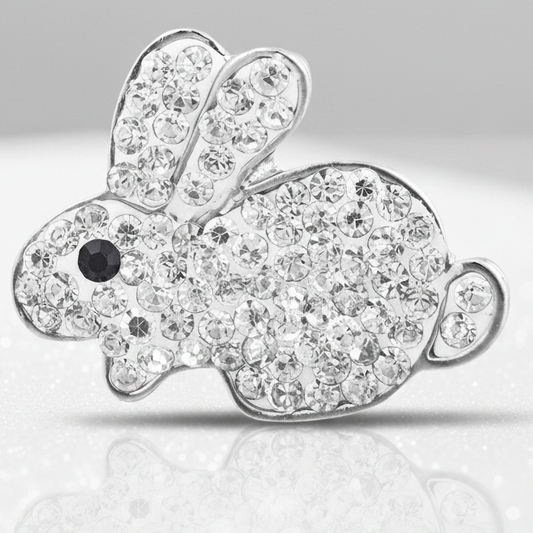 A silver bunny-shaped snap button covered in clear rhinestones, with a small black eye detail, displayed on a white surface with pastel Easter eggs and baskets in the background.