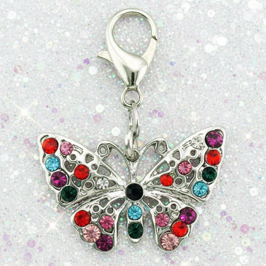 A silver butterfly-shaped charm embellished with colorful rhinestones in red, pink, blue, green, and black, attached to a silver lobster clasp, set against a sparkly white background.