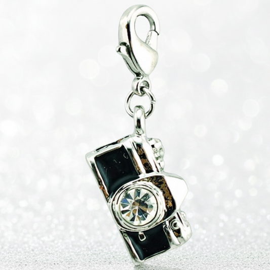 A silver charm shaped like a vintage camera with black enamel details and a clear rhinestone lens, attached to a silver lobster clasp, shown on a sparkly white background.